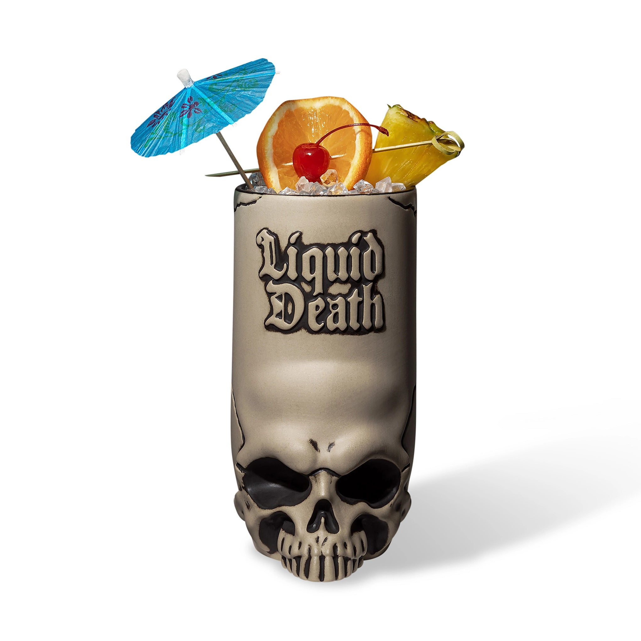 Skull-shaped cocktail glass with 'Liquid Death' text, filled with a tropical drink and garnishes on a white background.